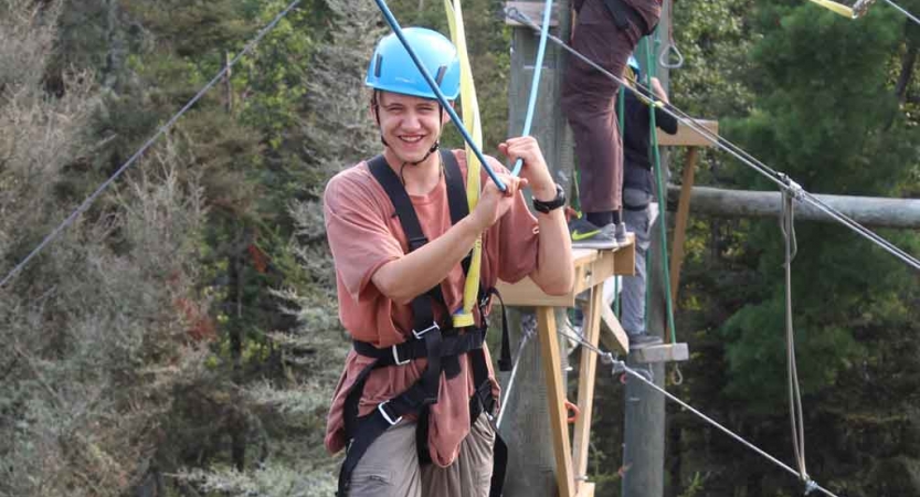 A person wearing safety gear and secured by ropes balances on an obstacle on a high ropes course. 